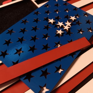 May include: A blue wooden cutout of the American flag with white stars. The flag is layered on top of red and white stripes.