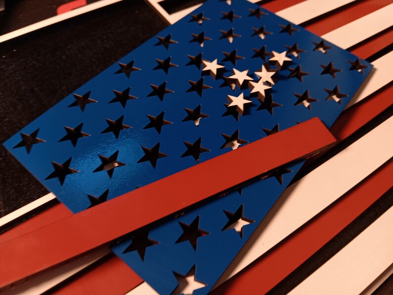 May include: A blue wooden cutout of the American flag with white stars. The flag is layered on top of red and white stripes.