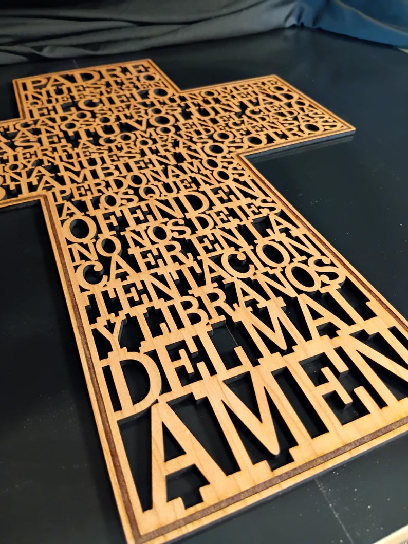 May include: A wooden cross with cut-out text in a script font. The cross is made of light-colored wood and the text is in Spanish. The text includes the words "Padre Nuestro" and "Amen".