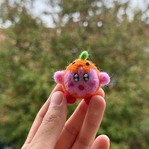 May include: A handmade felt character resembling a round, orange creature with pink details. It features small arms, feet, and a green stem. The character has two black eyes and is held in a person's hand.