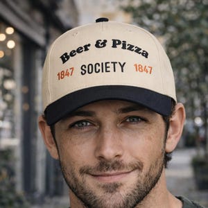 May include: A beige baseball cap with a black brim. The cap features the text "Beer & Pizza Society 1847" in black and orange lettering. The cap is worn by a person.