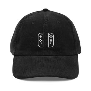 May include: Black corduroy baseball cap featuring a white embroidered design of two game controller halves. The cap has a curved brim and a button on top. A stylish accessory for gamers.