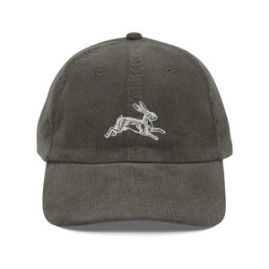 May include: Olive green corduroy baseball cap featuring a white embroidered running rabbit design. The cap has a curved brim and an adjustable strap at the back for a comfortable fit. A classic, casual accessory.