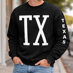 May include: Black crew neck sweatshirt with the white letters "TX" on the front. The word "TEXAS" is printed vertically in white on the right sleeve. The sweatshirt is worn with blue jeans.