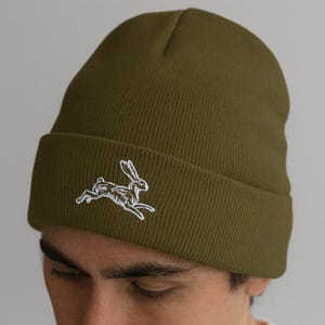 May include: Olive green beanie with a white embroidered running hare design. The hat is made of a soft, knitted material and has a folded brim. A stylish and comfortable accessory for cold weather.