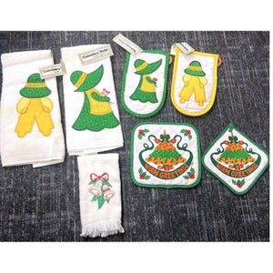 May include: A selection of white embroidered kitchen linens with festive designs. Includes towels, oven gloves, and a trivet. The designs feature a figure in a hat and overalls, and a Christmas tree with the words "Warm Greetings".