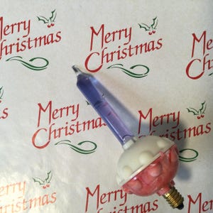 Może przedstawiać: Zabytkowa żarówka choinkowa z przezroczystą fioletową rurką i biało-czerwoną podstawą. Żarówka ma złotą podstawę śrubową. Tło zawiera słowa "Merry Christmas" w kolorze czerwonym.