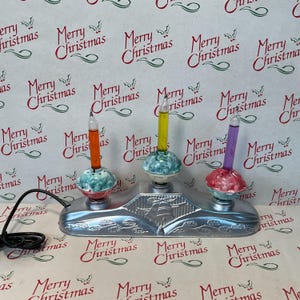 May include: A silver-toned electric holiday light fixture featuring three vertical bubble light tubes in orange, yellow, and purple. Each tube sits in a decorative base with a molded bell design on the front and a black power cord attached.
