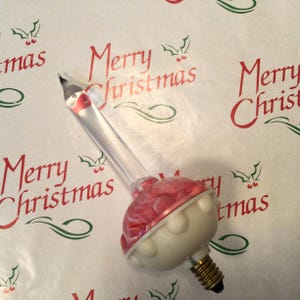 Może przedstawiać: Zabytkowa żarówka choinkowa z przezroczystą szklaną rurką i czerwoną i białą ozdobną podstawą. Żarówka ma mosiężną podstawę śrubową. Tło zawiera czerwone słowa "Merry Christmas".
