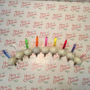 Op de afbeelding: Een verzameling vintage kerstlampen met kleurrijke kaarsvormige uiteinden. De witte lampen hebben een textuurontwerp, gerangschikt op een feestelijke achtergrond met "Merry Christmas"-patroon.