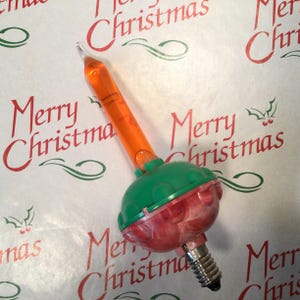 Może przedstawiać: Zabytkowa żarówka choinkowa z pomarańczową rurką i zielono-czerwoną podstawą. Żarówka ma przezroczystą końcówkę i podstawę wkręcaną. Tło to biały papier z powtarzającym się wzorem "Merry Christmas" w kolorze czerwonym.