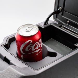 Może przedstawiać: Czerwona puszka Coca-Coli w uchwycie na kubek w konsoli samochodowej. Puszka z logo Coca-Coli jest na środku. Konsola jest ciemnoszara i ma pokrywę na zawiasach.