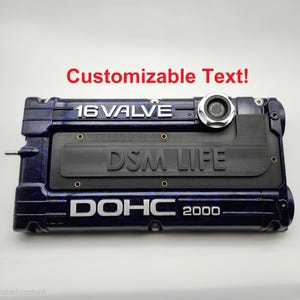 Może przedstawiać: Ciemnoniebieska pokrywa silnika z białym napisem "16 VALVE", "DSM LIFE" i "DOHC 2000". Pokrywa ma wzór tekstury i srebrny okrągły element. Tekst "Customizable Text!" jest w kolorze czerwonym.