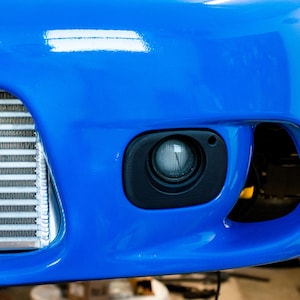 Może przedstawiać: Zbliżenie na jaskrawo niebieski zderzak samochodowy z czarną obudową światła przeciwmgielnego i srebrnym intercoolerem. Zderzak ma błyszczące wykończenie.