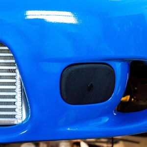 Może przedstawiać: Zbliżenie na jasnoniebieski zderzak samochodowy z czarną, prostokątną osłoną i dużym otworem. Srebrna i czarna chłodnica jest widoczna po lewej stronie zderzaka. Części samochodowe.