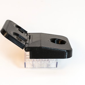 Könnte beinhalten: Eine schwarze, im 3D-Druck hergestellte, klappbare Box mit einem kleinen, durchsichtigen Kunststoffsockel. Die Box hat ein rundes Loch oben und zwei kleinere Löcher auf dem Deckel.