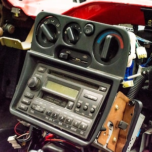 Może przedstawiać: Deska rozdzielcza samochodu z czarnym radiem i panelem sterowania klimatyzacją. Radio ma przyciski do CD, FM i kasety. Panel sterowania klimatyzacją ma pokrętła do regulacji temperatury i prędkości wentylatora. Widoczne są przewody i inne elementy.