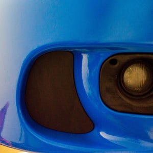 Może przedstawiać: Zbliżenie niebieskiego zderzaka samochodowego z czarną obudową światła przeciwmgielnego i czarnym akcentem. Światło przeciwmgielne ma przezroczystą soczewkę i żółtą żarówkę. Zderzak jest błyszczący.