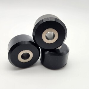 May include: Three black cylindrical objects with a central hole and a light-colored interior are stacked on a white background. The objects have a smooth, matte finish and appear to be made of a solid material.
