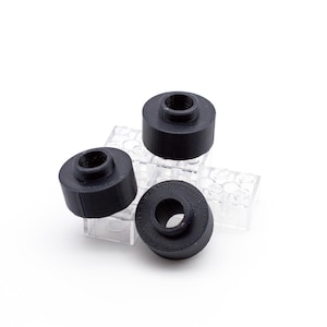 Może przedstawiać: Trzy czarne, cylindryczne obiekty z centralnym otworem ułożone są na przezroczystej, teksturowanej powierzchni. Obiekty mają zaokrąglony kształt i matowe wykończenie. Tło jest białe.