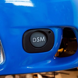Może przedstawiać: Zbliżenie na jasnoniebieski zderzak samochodu z czarnym okrągłym emblematem. Emblemat ma białe litery "DSM". Srebrna chłodnica jest widoczna po lewej stronie emblematu.