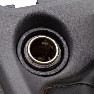 May include: Close-up of a black car interior component featuring a circular power outlet. The outlet has a silver-colored metal ring and is set within a textured, dark plastic panel. The image highlights the details of the automotive part.