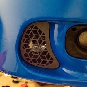 Może przedstawiać: Zbliżenie niebieskiego zderzaka samochodowego z czarnym geometrycznym otworem wentylacyjnym i okrągłym reflektorem. Otwór wentylacyjny ma wzór połączonych ze sobą rombów. Reflektor to ciemne koło.