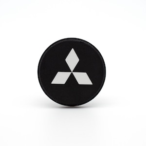 Może przedstawiać: Okrągła czarna naklejka z białym logo Mitsubishi. Logo to trójramienna gwiazda.