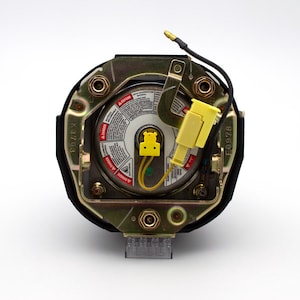 May include: Close-up of a black and gold steering wheel airbag module with a yellow connector and wiring. The module has a circular label with safety instructions and the text "E40974".