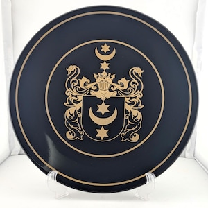 May include: A black ceramic plate with a gold-colored coat of arms featuring a crescent moon and stars. The plate is on a white stand.
