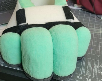 May include: A green and white foam hand with black fabric accents. The hand has four fingers and a thumb.
