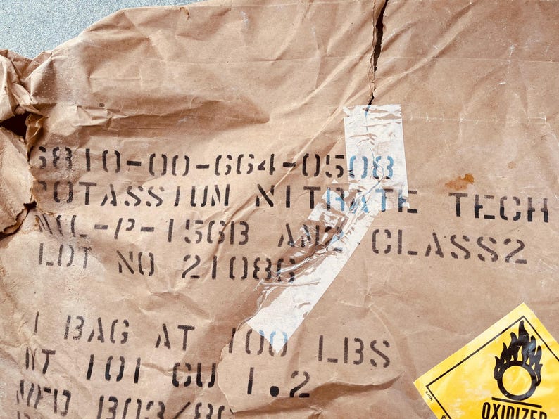 May include: A brown paper bag with black printed text that reads "6810-00-664-0503 POTASSIUM NITRATE TECH -12-15013 ANZ CLASS2 LOT NO 21086 1 BAG AT 100 LBS NT 101 CU 1.2 001303/80". A yellow and black warning label with the word "OXIDIZED" is visible on the right side of the bag.