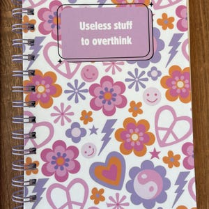 May include: A spiral-bound notebook with a colorful floral and graphic design. The cover features flowers, hearts, lightning bolts, and smiley faces in shades of pink, orange, and purple. The text "Useless stuff to overthink" is displayed on a lavender rectangle.