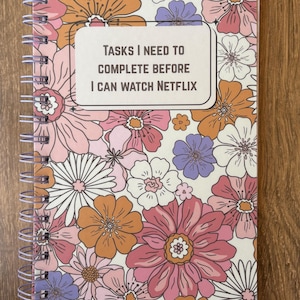 May include: Spiral-bound notebook with a floral design in shades of pink, orange, purple, and white. The cover features the text "TASKS I NEED TO COMPLETE BEFORE I CAN WATCH NETFLIX" in a white rectangle.