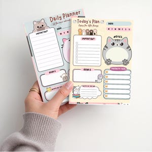 May include: Two daily planner notepads featuring cat illustrations. The left pad is light blue with the text Daily Planner. The right pad is cream-colored with the text Today's Plan, featuring sections for important tasks, goals, notes, and a priority task list.