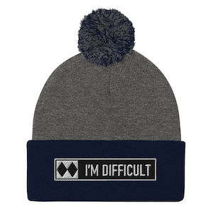 May include: Grey and navy blue knitted beanie with a black patch that says "I'M DIFFICULT" and a black diamond symbol. The beanie has a grey pom pom on top.