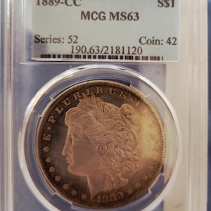 May include: An 1889-CC Morgan silver dollar coin encapsulated in a clear plastic slab. The label reads 1889-CC, S$1, MCG MS63, Series: 52, Coin: 42, and 190.63/2181120. The coin features a profile of Lady Liberty with the text E PLURIBUS UNUM.