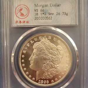 Può includere: Una moneta Morgan Dollar del 1895-O classificata MS 66, racchiusa in una custodia protettiva in plastica trasparente. La moneta presenta il profilo di Lady Liberty, il testo E PLURIBUS UNUM e l'anno 1895. L'etichetta indica 38,1 mm e 26,73 g.