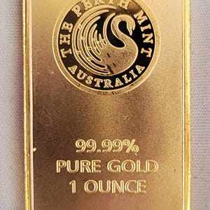 May include: A rectangular gold bullion bar featuring the Perth Mint Australia logo with a swan emblem. The text reads 99.99% PURE GOLD 1 OUNCE. The surface has a metallic finish with raised lettering and a circular design element at the top center.