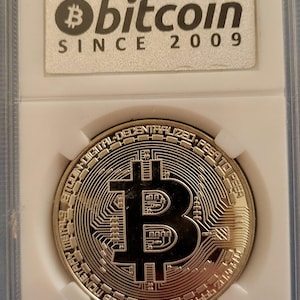 May include: A silver-colored Bitcoin coin encased in a rectangular plastic protective holder. The coin features a central Bitcoin symbol with circuit board patterns and text reading Bitcoin Digital Decentralized Peer to Peer. The label reads Bitcoin Since 2009.