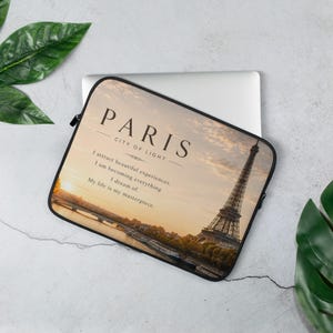 May include: A rectangular laptop sleeve featuring a sunset photograph of the Eiffel Tower in Paris. The text reads PARIS CITY OF LIGHT followed by I attract beautiful experiences. I am becoming everything I dream of. My life is my masterpiece.