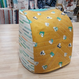 May include: A quilted fabric cover for a sewing machine. The cover is yellow with a pattern of sewing machines and has a teal and white striped side.