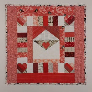 May include: A red, white, and pink patchwork quilt with a heart-shaped envelope design. The quilt is framed with a pink and white floral border.