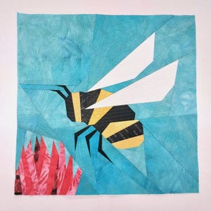 May include: A square fabric quilt block featuring a pieced bee design. The bee has black and yellow striped segments and white wings. The background is composed of various shades of teal fabric, with a pink floral element in the bottom left corner.