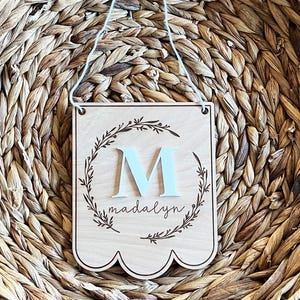 May include: A wooden sign with a scalloped edge, featuring a light blue letter "M" surrounded by a wreath of leaves. The name "Madalyn" is written below the letter.