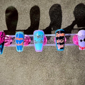 May include: Five colourful fake fingernails with different designs. The designs include a pink and orange swirl, a blue and orange design with the word "worm" written on it, a blue and orange design with the words "Frog's Breath" written on it, a blue and orange design with the words "Deadly Night Shade" written on it, and a white design with a black face and sunglasses.