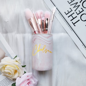 Op de afbeelding: Een roze en witte marmeren make-up kwastenhouder met de naam "Chelsea" in gouden letters.