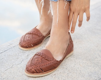 May include: A pair of brown leather sandals with woven straps. The sandals are worn by a person wearing blue jeans.