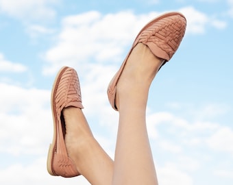 May include: A pair of light brown leather sandals with woven straps. The sandals are worn by a person with their legs raised in the air against a blue sky with white clouds.