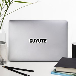 Puede incluir: Un ordenador portátil plateado con una pegatina negra que dice "GUYUTE" en la tapa. El portátil está sobre una superficie blanca con una taza blanca, dos lápices y una pila de libros.